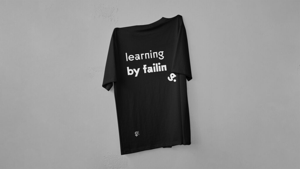 Na zdjęciu znajduje się czarna koszulka wisząca na ścianie. Na jej plecach widnieje napis: "Learning by failing". W dolnym rogu koszulki znajduje się niewielkie logo kierunki Grafika.